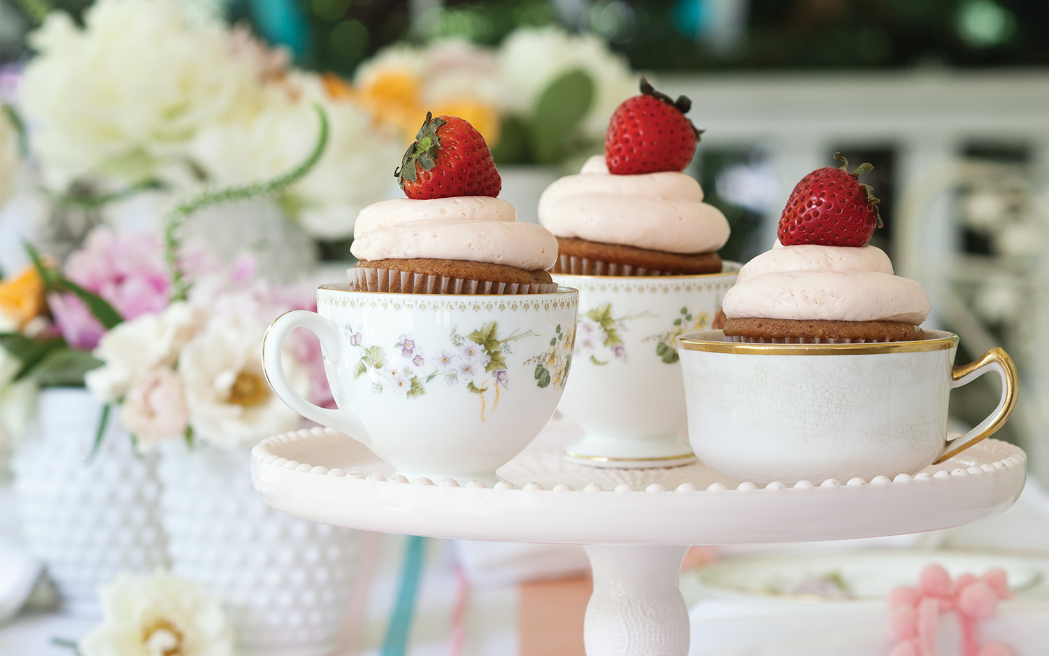 Three strawberry cupcakes in mismatched teacups on a cake stand