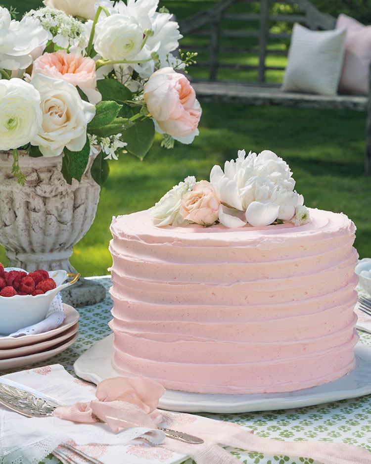 A tall pink-frosted cake topped with fresh flowers that match a nearby arrangement