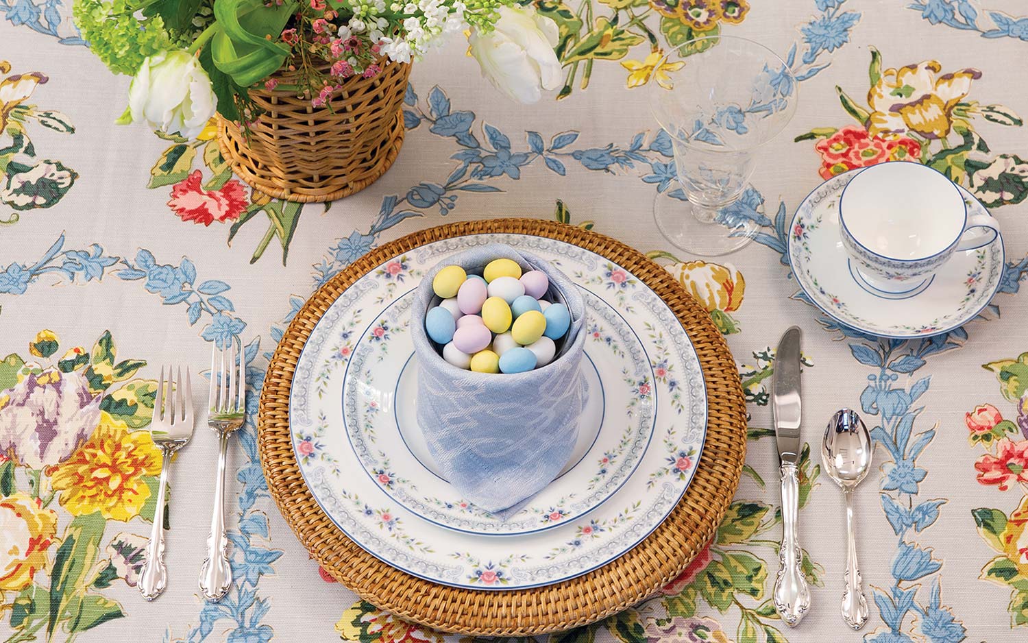 Floral tablecloth, woven place mat, china setting adorned with pink and blue flowers showcasing a cupful of pastel candies wrapped in a pale blue napkin