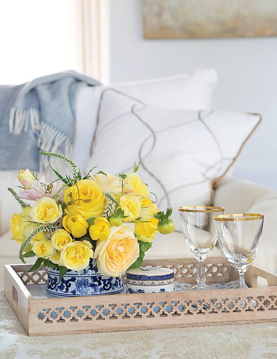 A vignette composed of yellow flowers in a blue-and-white container and two gold-rimmed wine glasses on a coffee table tray