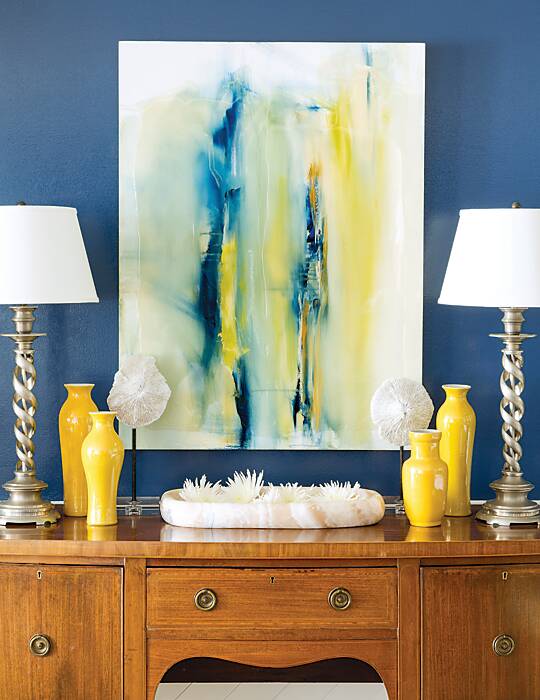 Blue, yellow, and white abstract painting above a wood sideboard and flanked by yellow vases and silver table lamps