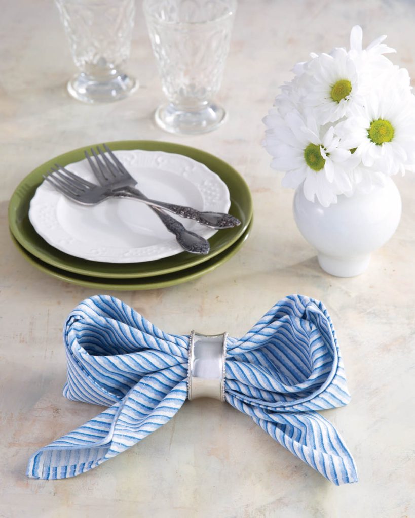 Napkin Fold How-To: A blue-and-white napkin folded into a bow, white flowers in a vase, a stack of white and green plates topped with silverware