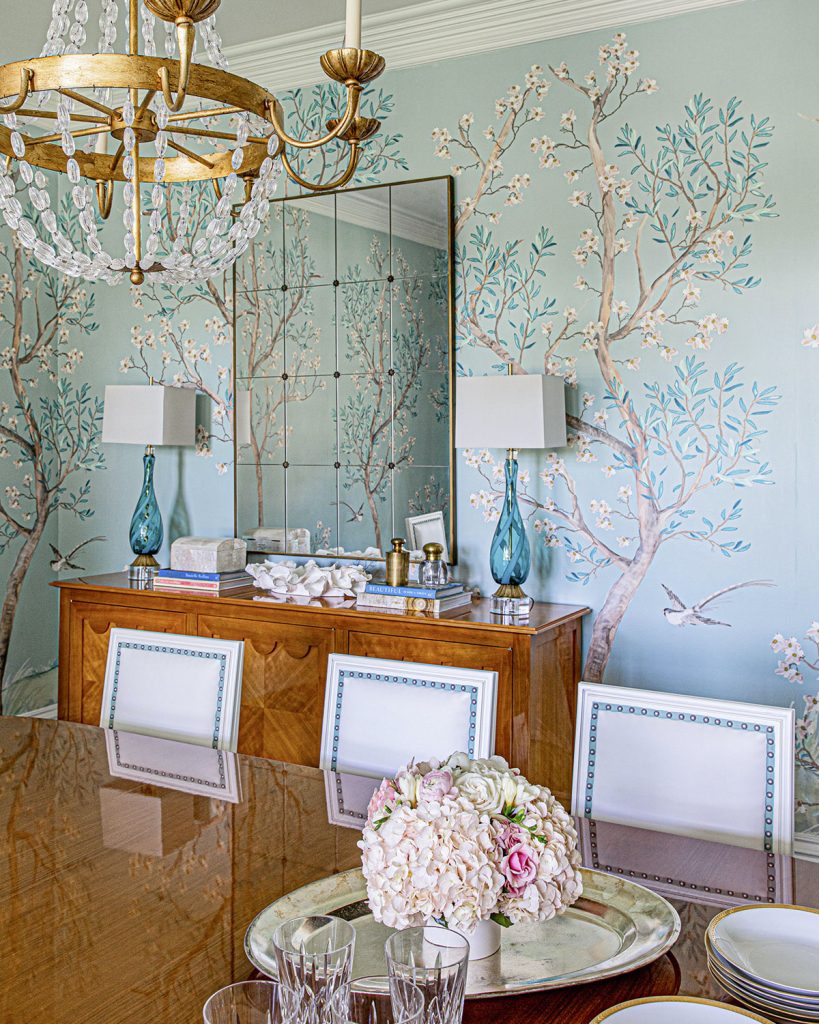 Dining room with blue garden scene wallpaper, light-colored high-polish furnishings, and a gold beaded chandlier