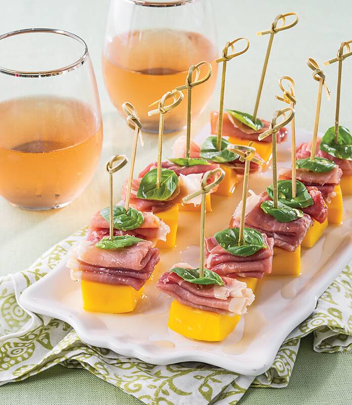 Mango, prosciutto, and basil leaves on small bamboo skewers on a white platter