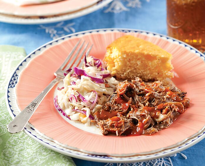 A pale pink plate features sauced pulled pork, coleslaw, cornbread, and a silver fork